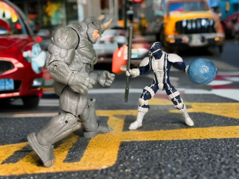 Boneco do Cardiac segurando seu bastão e uma explosão de energia azul, enfrentando o boneco do Rhino em uma rua.