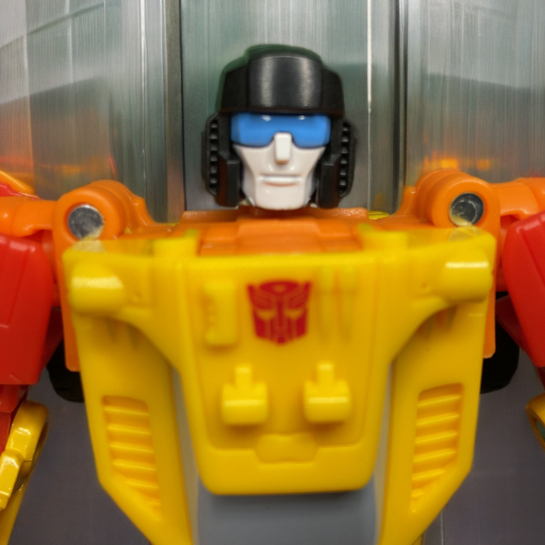 Detalhe em close da cabeça do Sureshot no modo robô, capacete preto, placa facial prateada e visor azul brilhante.
