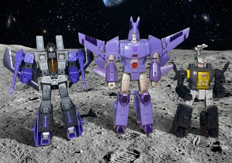 Tres figuras de acción Transformers, Skywarp, Cyclonus y Bombshell, posan en fila sobre la superficie rocosa de una luna bajo un cielo estrellado del espacio profundo.