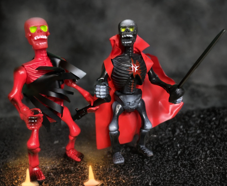 A close-up studio photograph of two 5.5-inch vintage skeleton action figures standing shoulder-to-shoulder on a dark, rocky surface. On the left is Ribs, an off-white skeletal figure with an exposed ribcage and tattered grey fabric vest remnants. On the right stands Crossbones, featuring a distinct skull head-sculpt, vibrant red with a white skull-and-crossbones emblem, and black leg greaves. Both figures have their classic five-point articulation joints posed symmetrically.