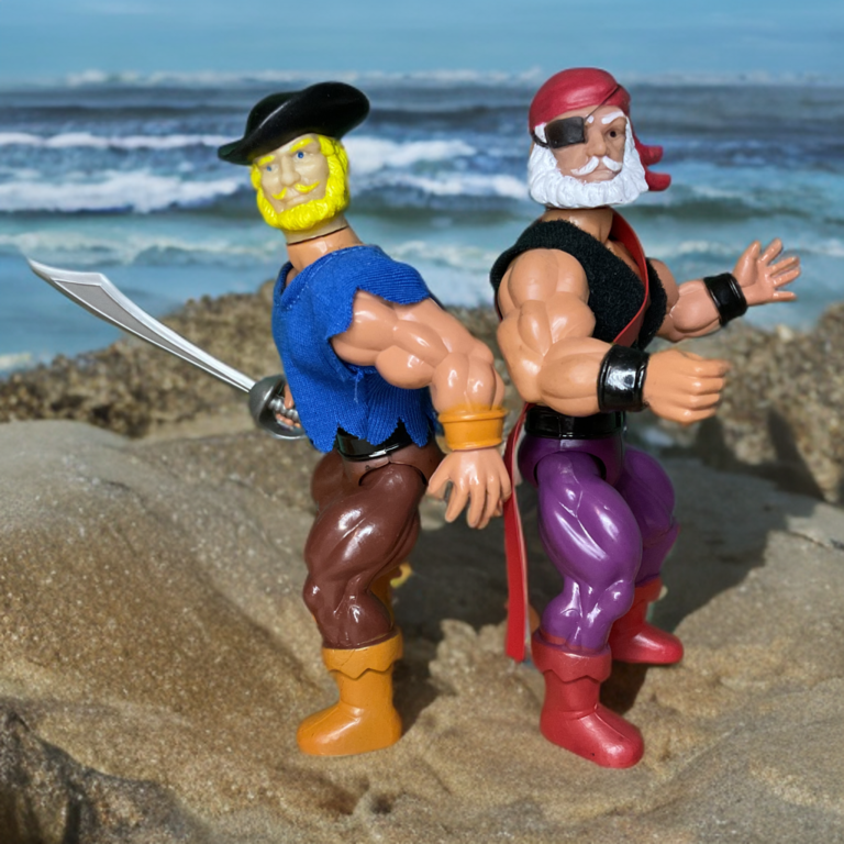 A close-up, ground-level outdoor photograph of two vintage 5.5-inch action figures standing side-by-side on wet beach sand. On the left is Cutlass, an athletic figure with long brown hair, a painted black mustache, a bright red-and-gold layered sash, and purple trousers. On the right is Patch, sporting his iconic molded red bandana, a classic black eye patch over his right eye, and a blue vest. In the background, blue ocean waves roll onto the shore beneath a bright, clear sky.