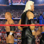 A close-up shot of four professional wrestling action figures standing inside a miniature wrestling ring with a red ring rope running across the foreground. On the left side of the ring, an action figure of Road Warrior Hawk in blue and black face paint and spiked shoulder pads stands alongside a classic blond wrestler figure in black trunks. They face off against two figures on the right side of the ring in this pre match standoff.
