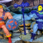 Vintage 1982 Masters of the Universe He-Man and Skeletor action figures standing on a rocky terrain in front of Castle Grayskull. He-Man is on the left, holding a silver half-sword overhead and wielding a grey and red shield. Skeletor is on the right, wearing his signature purple hood and chest armor while holding his dark purple Havoc Staff.